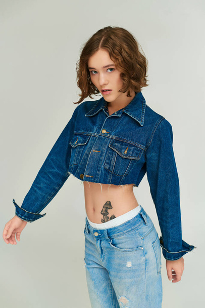 fashionable girl with tattoo in cropped denim jacket and blue jeans posing on grey backdrop - Photo, Image