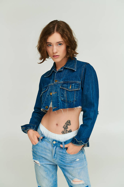 симпатичная девушка в модном джинсовом пиджаке, позирует с руками в карманах джинсов на сером фоне - Фото, изображение