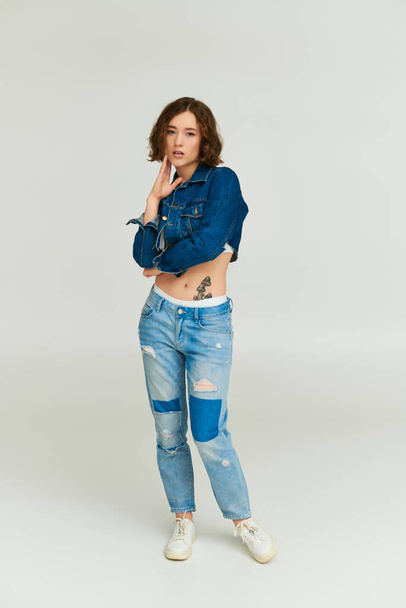 stylish young model with tattoo in cropped denim jacket and blue jeans posing on grey backdrop - Photo, Image