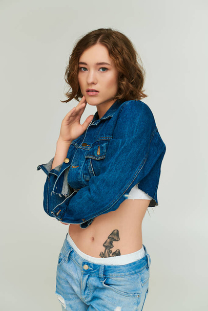 modelo joven de moda con tatuaje en chaqueta de mezclilla recortada y vaqueros azules posando sobre fondo gris - Foto, imagen