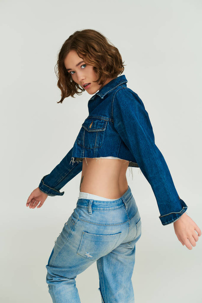 fashionable young model in cropped denim jacket and blue jeans walking on grey background - Photo, Image