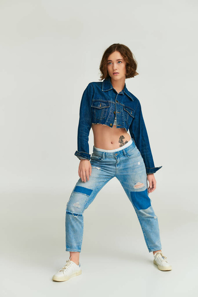 zelfexpressie, trendy jong model in bijgesneden spijkerjasje en blauwe jeans poserend op grijze achtergrond - Foto, afbeelding