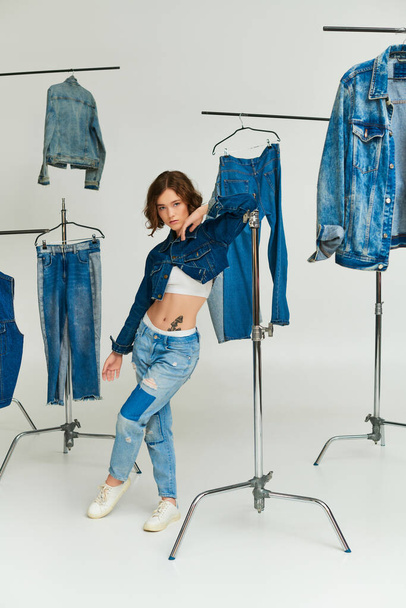 stylish model with tattoo posing in cropped jacket and jeans among denim clothes on grey backdrop - Photo, Image