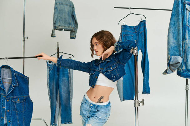stylowy model z tatuażem pozujący w obciętej kurtce i dżinsach wśród odzieży jeansowej na szarym tle - Zdjęcie, obraz