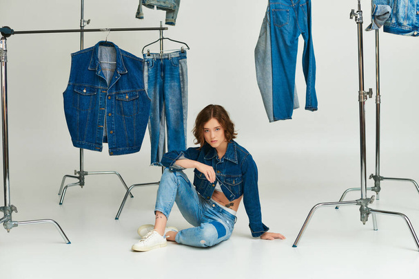 stylisches Model mit Tätowierung posiert in abgeschnittener Jacke und Jeans sitzend zwischen Jeanskleidung auf grau - Foto, Bild