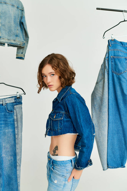 stylische junge Frau mit Tätowierung in abgeschnittener Jacke und Jeans, die zwischen Jeanskleidung auf grau steht - Foto, Bild