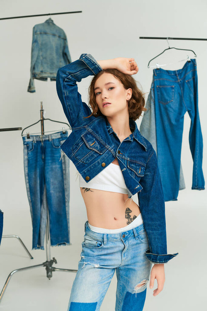 tattooed young model in cropped jacket and jeans posing with raised hand among denim clothes on grey - Photo, Image
