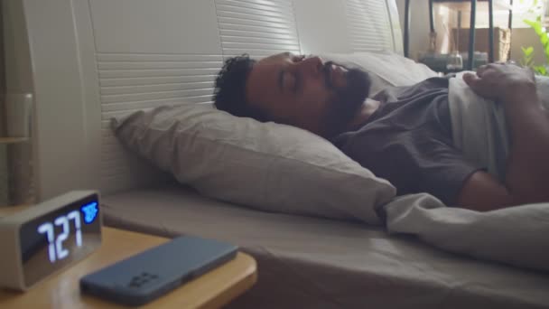 Fiatal szakállas férfi felébred az ágyban, kikapcsolja az ébresztőórát és okostelefont használ reggel otthon. - Felvétel, videó