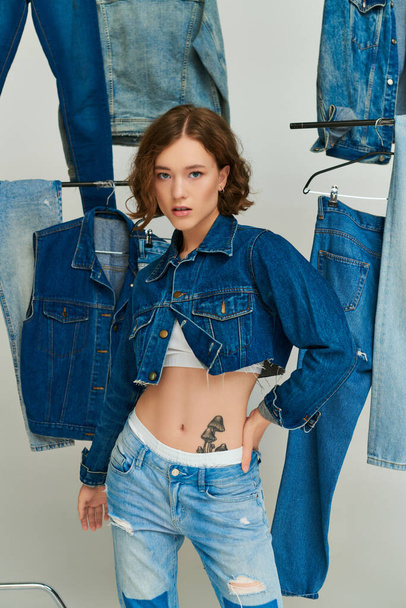красивая девушка в обрезанной куртке и джинсах позирует с рукой на бедре среди модной джинсовой одежды на сером - Фото, изображение