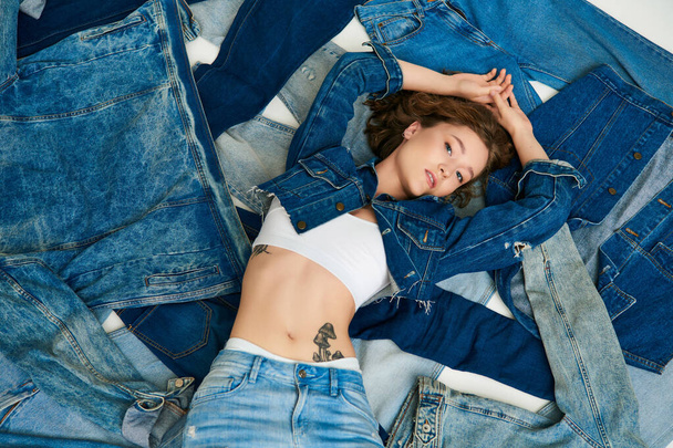 вид сверху, татуированная молодая модель лежащая на куче джинсовой одежды с поднятыми руками, быстрая мода - Фото, изображение