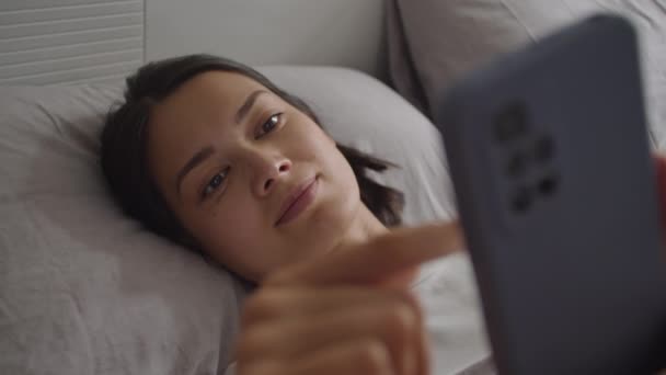 Mujer joven acostada en la cama y desplazándose a través de las redes sociales en el teléfono después de despertarse por la mañana en casa - Imágenes, Vídeo