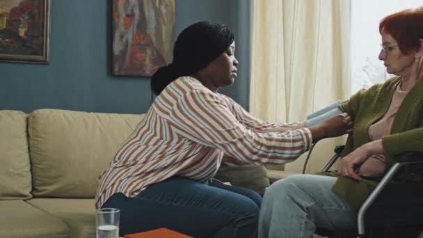 Panorámica derecha de mujer mayor sentada en silla de ruedas y enfermera afroamericana revisando su presión arterial en la sala de estar durante el día - Imágenes, Vídeo