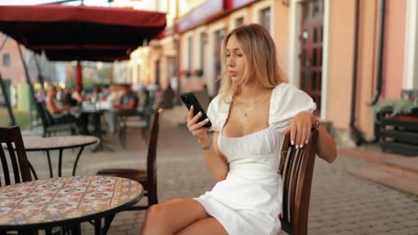 Ritratto ravvicinato di una bella giovane donna bionda con occhi affascinanti che tiene in mano uno smartphone, seduta a un tavolo in un caffè di strada e sorridente dolcemente all'aperto - Filmati, video