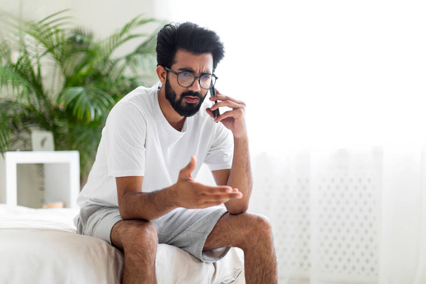 Portrait Of Worried Young Indian Man Talking On Cellphone In Bedroom, Upset Stressed Eastern Guy Sitting On Bed And Having Unpleasant Phone Conversation, Closeup Shot With Copy Space - Photo, image