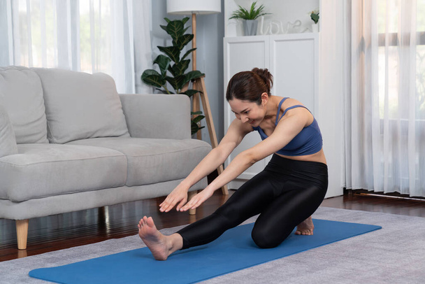 Mujer asiática en ropa deportiva haciendo ejercicio de yoga en la alfombra de fitness como su rutina de entrenamiento en casa. Cuidado corporal saludable y meditación tranquila en el estilo de vida del yoga con comodidad y relajación. Vigoroso - Foto, Imagen