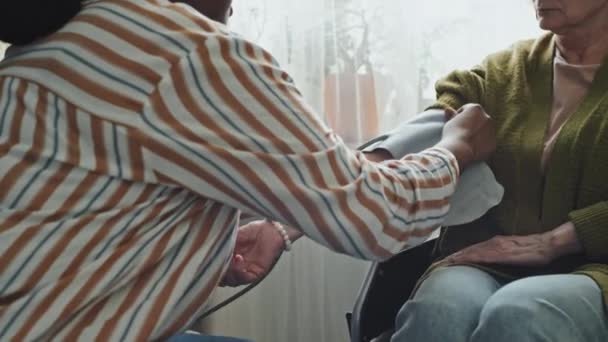 Inclinazione di donna anziana preoccupata in sedia a rotelle e assistente di assistenza domiciliare afroamericana che controlla la pressione sanguigna con il pulsometro in soggiorno durante il giorno - Filmati, video