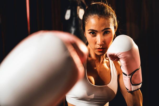 Asiatische Muay Thai Boxerin schlägt vor der Kamera die Faust in kampfbereiter Haltung, posiert im Fitness-Studio mit Boxausrüstung im Hintergrund. Konzentrierte Entschlossenheit und Vorbereitung auf die Herausforderung. Impulse - Foto, Bild
