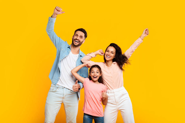 Victory concept. Overjoyed european family of three shaking clenched fists, making winner gesture, posing over yellow background. Father, mother and their daughter celebrating success together - Photo, Image
