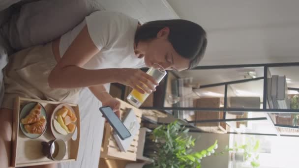 Вертикальний знімок високого кута молодої жінки, яка снідає в ліжку, п'є апельсиновий сік, їсть фрукти і прокручується на сенсорному екрані смартфона вранці вдома - Кадри, відео