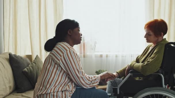 Panning prawy średni strzał zdenerwowanej starszej kobiety na wózku inwalidzkim i Afroamerykańskiej asystentki domu uspokajając ją i trzymając ją za rękę w salonie w ciągu dnia - Materiał filmowy, wideo
