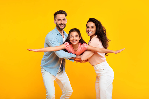 Cheerful european parents playing with their daughter holding girl in arms posing on yellow background and smiling at camera. Family having fun together, kid spreading hands like plane - Photo, Image