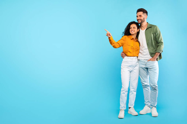 Για δες αυτό. Χαρούμενο ευρωπαϊκό ζευγάρι που επιδεικνύει το χώρο αντιγραφής για τη διαφήμισή σας, γυναίκα που δείχνει κατά μέρος τον ελεύθερο χώρο σε μπλε φόντο. Πλήρες στούντιο πλάνο των ευτυχισμένων συζύγων - Φωτογραφία, εικόνα