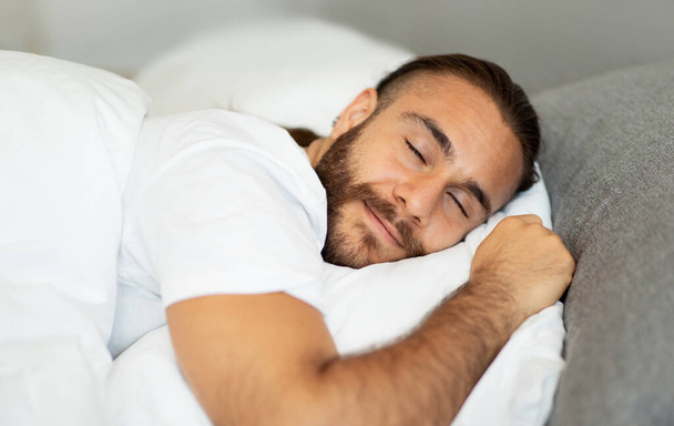 Cómodo concepto de ambiente saludable para dormir. Foto de primer plano de pacífico guapo barbudo hombre milenario en camiseta blanca durmiendo en la cama en casa, acostado en el vientre, sonriendo - Foto, Imagen