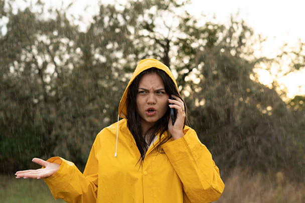 Недовольная молодая женщина использует мобильный телефон, чтобы звонить во время дождя. Вызвать такси в дождливую погоду. Копирование пространства - Фото, изображение