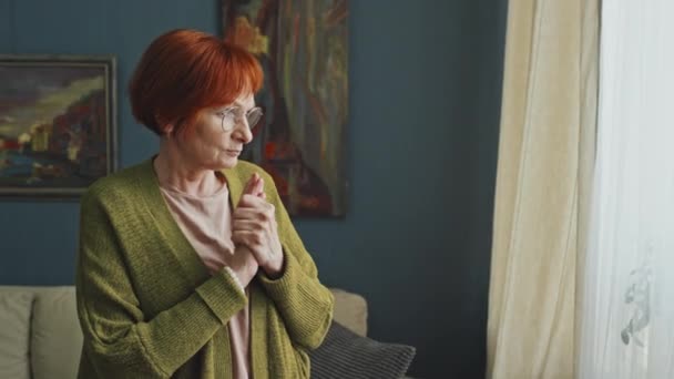 Keskikokoinen laukaus melankolinen vanhempi nainen seisoo ikkunan vieressä olohuoneessa ja harkitsee etäisyyttä - Materiaali, video