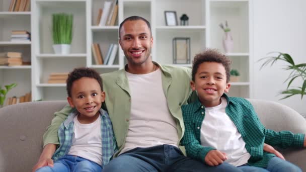 Πορτρέτο του χαρούμενου πατέρα αγκαλιάζει δύο χαμογελαστούς γιους ενώ χαλαρώνει σε μαλακό καναπέ στο σύγχρονο σαλόνι. Ευτυχισμένη πολυεθνική οικογένεια σε casual ρούχα απολαμβάνοντας το χόμπι μαζί στην άνεση του σπιτιού. - Πλάνα, βίντεο