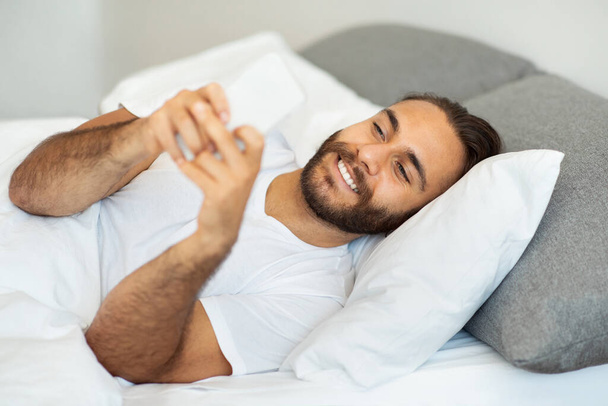 Pozitivní atraktivní vousatý mladík v bílém tričku leží ráno v posteli, drží mobil, dívá se na obrazovku a usmívá se. Happy man reading news, scrolling on social media, chat with lover - Fotografie, Obrázek