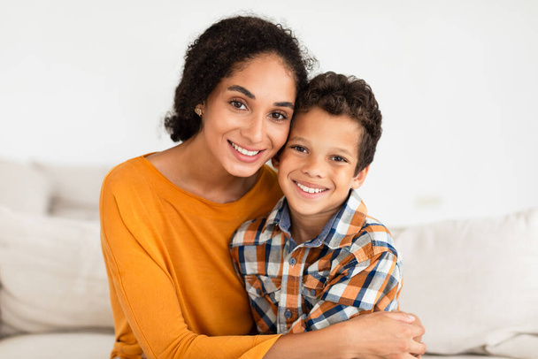 Portret van een gelukkige jonge moeder omhelst haar zoon glimlachend naar camera thuis Interieur. Jongen geniet van moeders knuffel zittend op de bank binnen. Familie binding en moederschap concept - Foto, afbeelding