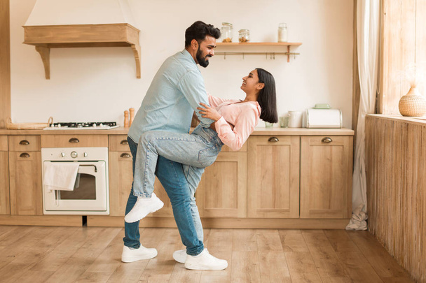 Romantic indian couple having fun together, feeling happy, dancing in kitchen interior, full length, side view. Joy, romantic date, celebration anniversary and positive weekend at home - Photo, Image