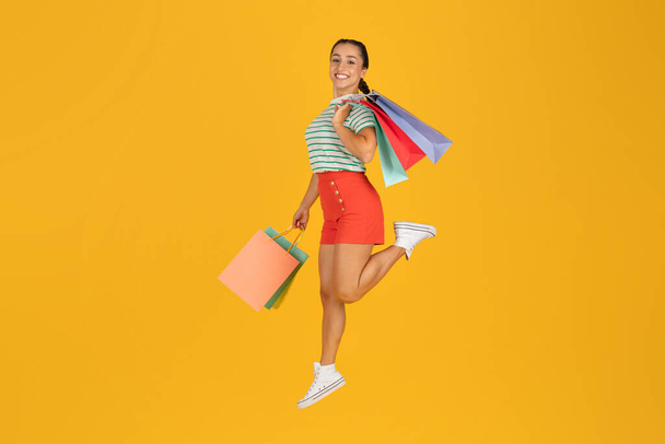 Positiv glückliche attraktive junge Frau in stylischem Outfit shopaholic mit bunten Einkaufstaschen in den Händen springen in die Luft auf gelbem Hintergrund, volle Länge, Kopierraum. Verkaufssaison, schwarzer Freitag - Foto, Bild