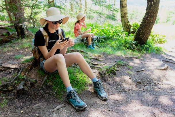 Młoda dziewczyna podróżnik siedzi na ziemi w lesie i za pomocą smartfona. - Zdjęcie, obraz