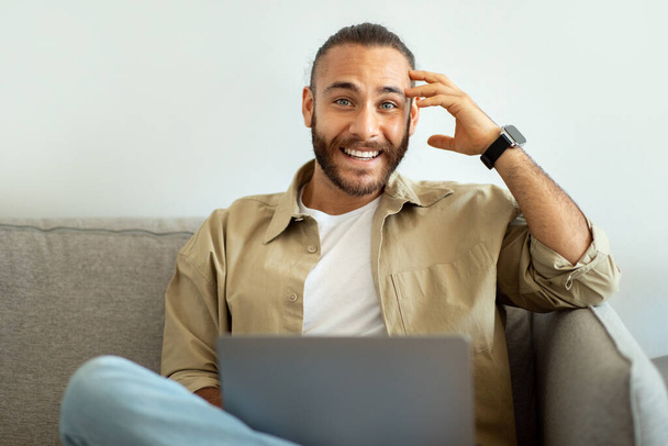 Nadšený šťastný hezký mladý muž s chytrými hodinkami na zápěstí sedí na pohovce, pomocí notebooku, na volné noze pracuje z domova, kopírovat prostor pro vzdálenou práci reklamy - Fotografie, Obrázek