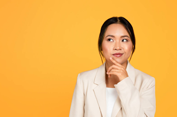 Pensivo positivo giovane donna asiatica in giacca e cravatta pensa, guardando lo spazio vuoto, isolato su sfondo giallo studio. Scelta, emozioni della gente, annuncio e offerta, soluzione e idea creano - Foto, immagini