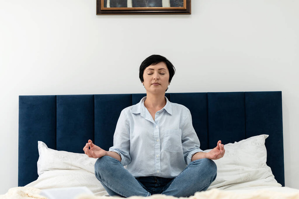 Brunette middle aged woman in a shirt and jeans doing yoga meditation at home in the bedroom, mental health. Peaceful woman meditate to achieve mental balance, time for herself at home. - Photo, Image