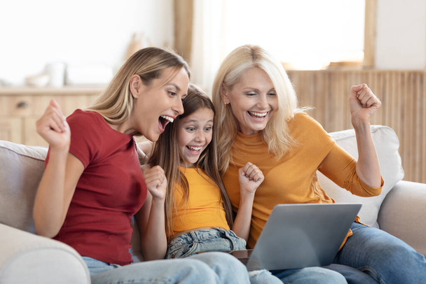 Emotional happy beautiful blonde grandmother, mom and girl kid daughter using notebook computer at home, looking at pc screen, clenching fists and exclaiming, shopping online, got cashback - Photo, Image