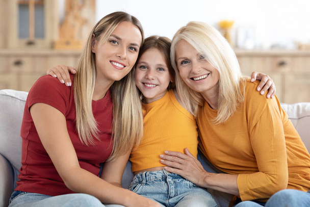 Family portrait of happy beautiful blonde loving mother, grandmother and child preteen girl at home. Cute child daughter hugging her mom and granny, posing on couch for family picture - Photo, Image