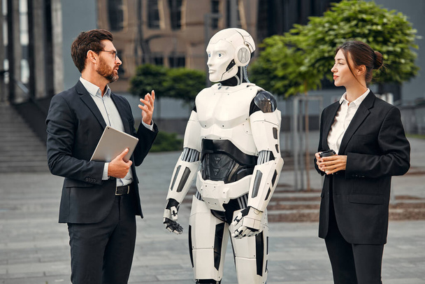 Молодой человек и женщина деловые партнеры в костюмах с планшетом, стоящие вместе с роботом и обсуждающие работу на фоне здания. Будущее с искусственным интеллектом. - Фото, изображение