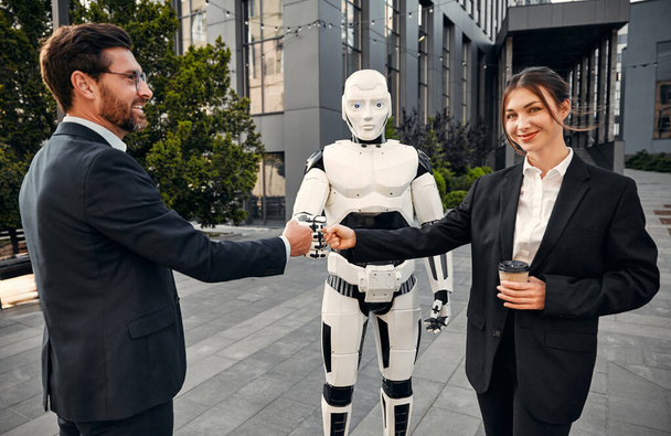 Young man and woman business partners in suits standing together with a robot and connecting their fists together. The future with artificial intelligence. - Photo, Image
