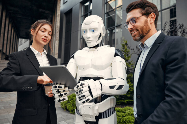 Νέοι άνδρες και γυναίκες επιχειρηματικοί συνεργάτες με κοστούμια με ένα tablet που στέκονται μαζί με ένα ρομπότ και συζητούν για την εργασία στο φόντο ενός κτιρίου. Το μέλλον με την τεχνητή νοημοσύνη. - Φωτογραφία, εικόνα