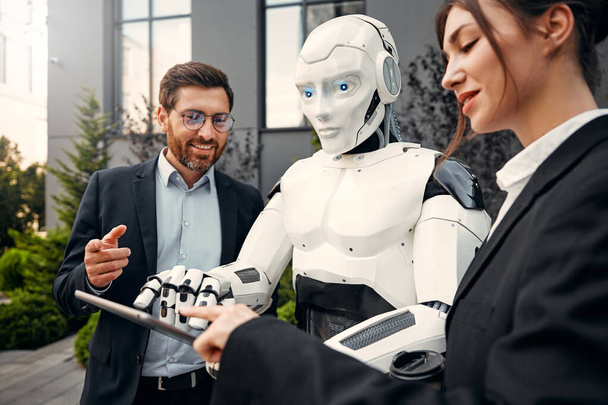 Jeunes hommes et femmes partenaires d'affaires en costumes avec une tablette debout avec un robot et discuter du travail dans le contexte d'un bâtiment. L'avenir avec l'intelligence artificielle. - Photo, image
