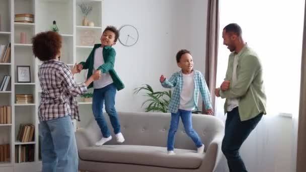 Grappige actieve familie van Afrikaanse Amerikaanse etniciteit doen feest en dansen samen in de stijlvolle woonkamer. zorgeloze kleine kinderen springen blootsvoets op de bank en plezier hebben met mama en papa thuis. - Video