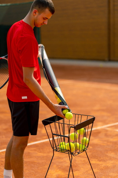 крупным планом теннисист собирает мячи с трубкой на открытом оранжевом теннисном корте спортивного инвентаря - Фото, изображение