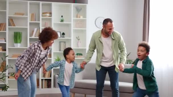Dört çocuklu Afro-Amerikan ailesi evde harika vakit geçiriyor. Neşeli ebeveynler ve sıradan giyinmiş iki sevimli çocuk el ele tutuşup geniş oturma odasında dans ediyorlar.. - Video, Çekim