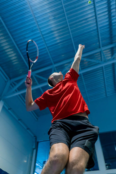 junger Mann auf einem überdachten Tennisplatz schlägt den Ball mit einem Schläger in einem Sprungaufschlag während des Spiels Tennislehrer Profisport - Foto, Bild