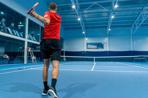 junger Mann auf einem überdachten Tennisplatz schlägt den Ball mit einem Schläger servieren während des Spiels Tennislehrer Profisport - Foto, Bild