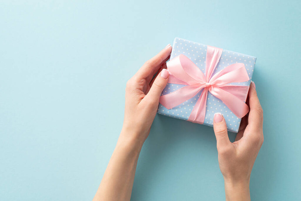 El arte de regalar: Desde la vista superior en primera persona, la niña presenta una caja de regalo azul pastel adornada con lunares y un lazo de cinta rosa, contra un fondo azul pastel para su toque personal - Foto, imagen